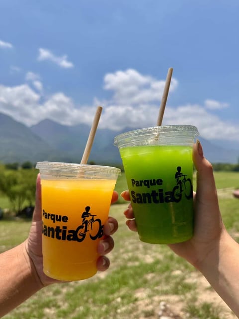 Two hands holding colorful beverages with Parque Santi logo against mountain landscape background