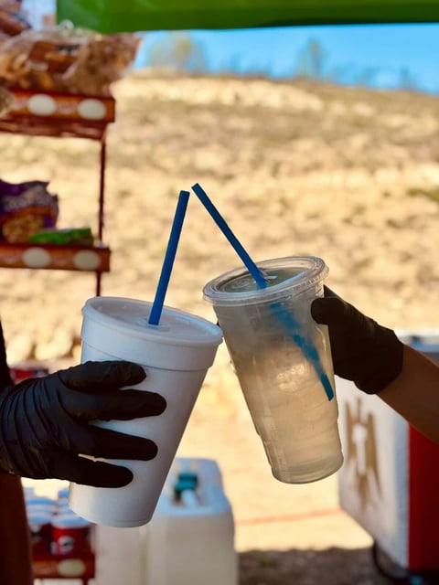 Person in black gloves holding two clear plastic cups with blue straws against a desert landscape