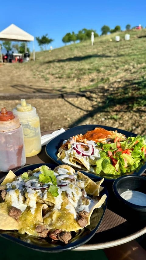Outdoor picnic on a hillside with loaded nachos, salad, drinks, and a white tent canopy in the background under clear blue sky